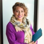 Smiling woman in a purple dress and scarf holding a tablet indoors.